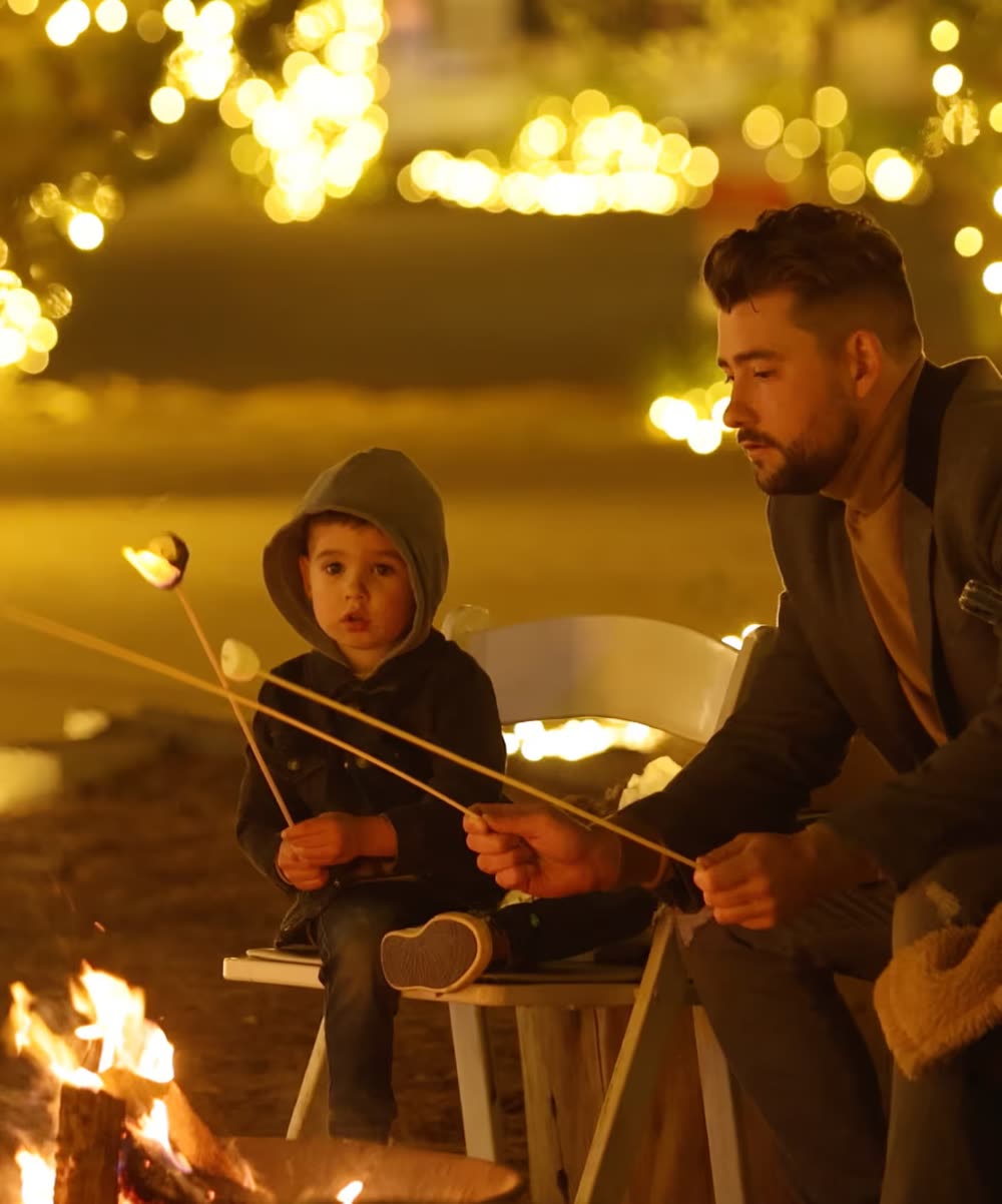 A father and young son roasting marshmallows by a fire under glowing lights